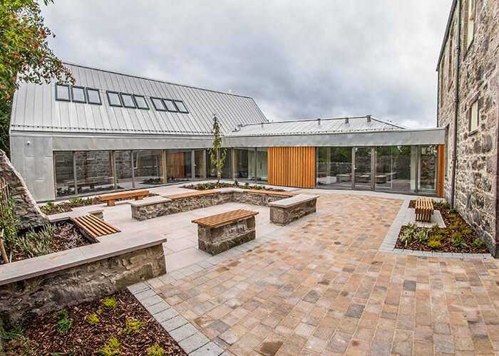 View of court-yard at Kingussie Courthouse. Outside seating area and gardens with building extension with large windows.
